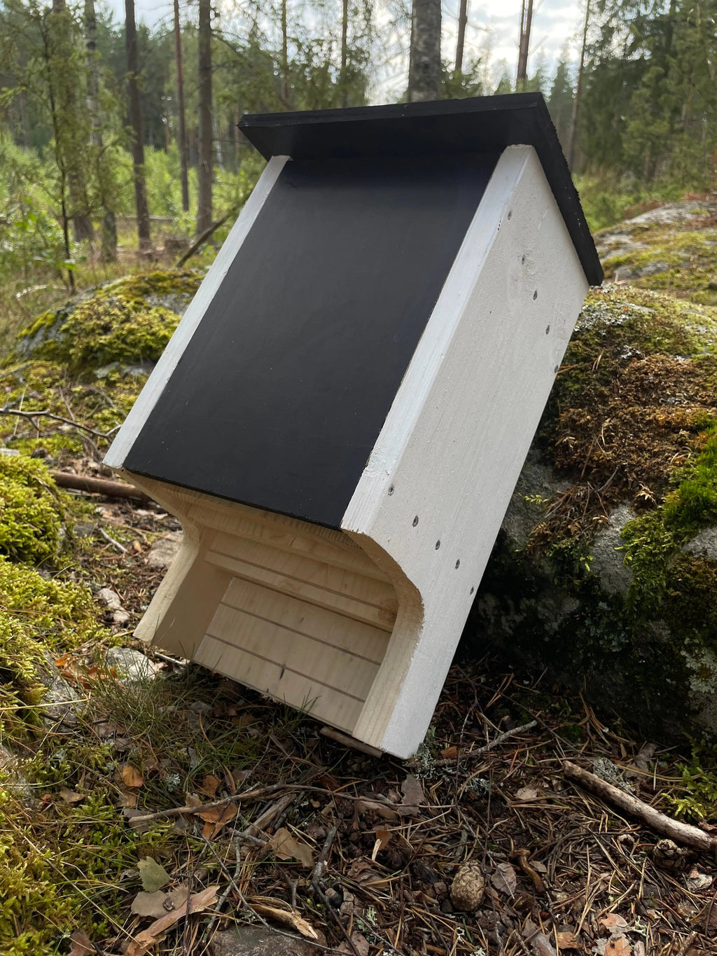 Vit målad fladdermusholk i trä lutad mot sten i grönområde som holk för fladdermöss. framsida, tak, ingångshål och högersida synligt. 